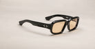 The Jacques Marie Mage Nakahira sunglasses feature black rectangular thick frames, light brown lenses, and temples adorned with small white decorative dots, displayed on a plain beige background.