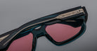 Close-up of Jacques Marie Mage Nakahira sunglasses featuring dark blue-green frames, pink-tinted lenses, and an ornate metallic temple design, set against a neutral gray background.