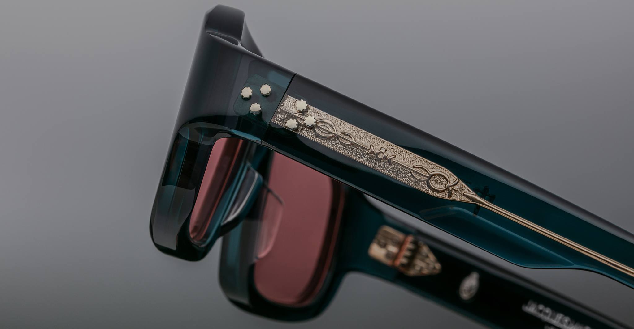 Close-up of two Jacques Marie Mage Nakahira sunglasses with dark frames and pink lenses, featuring intricate gold details and small star motifs on the arms, set against a blurred grey background.