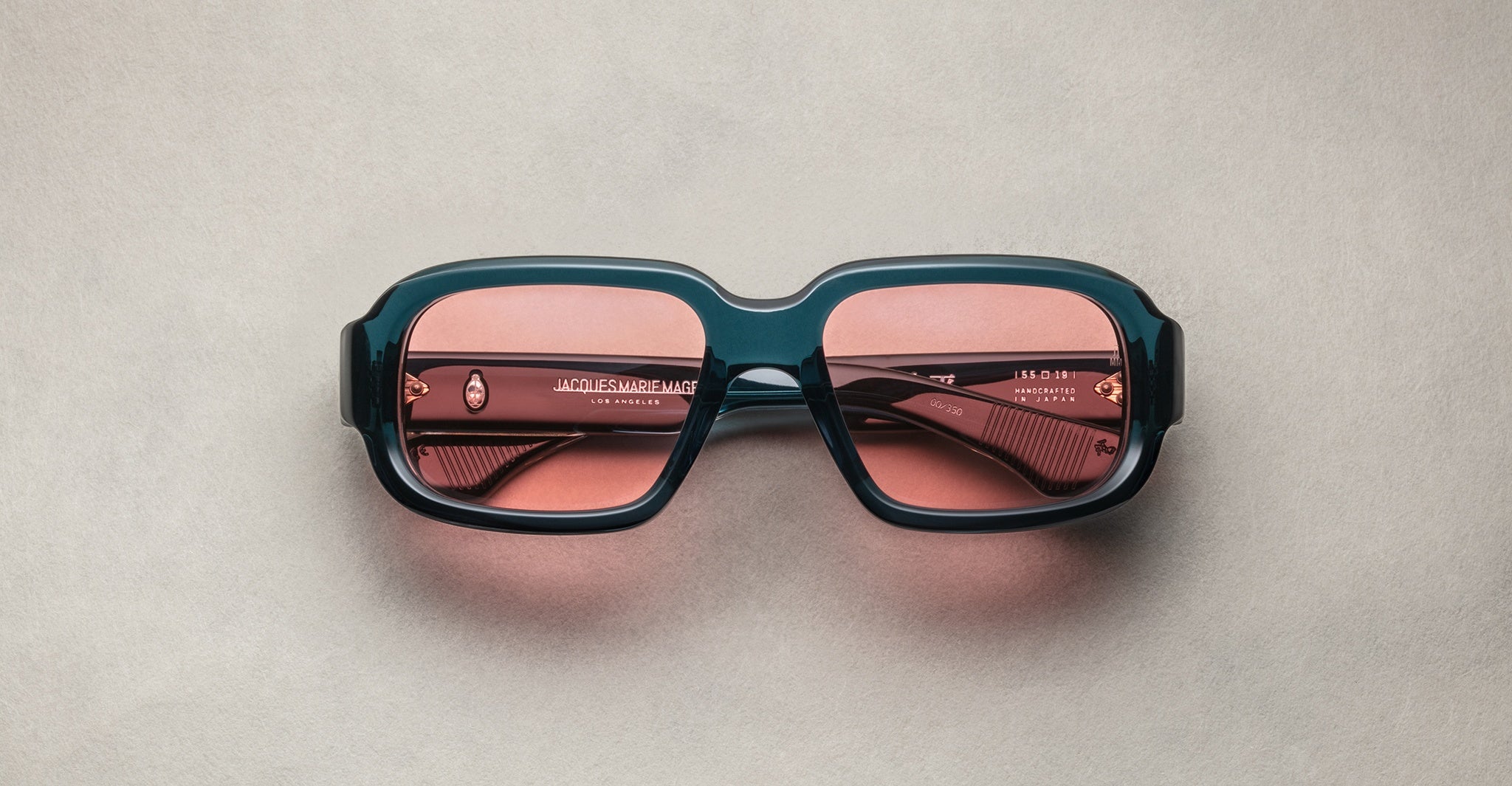 The Jacques Marie Mage Nakahira sunglasses, featuring chunky rectangular teal frames and pink-tinted lenses, are displayed front-facing with slightly closed arms on a plain light gray surface.