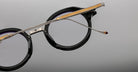 Jacques Marie Mage Norman eyeglasses, black round frame with gold and silver arms, front view.