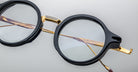 Jacques Marie Mage Norman round eyeglasses, black and gold frames, clear lenses, front view.