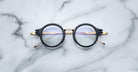 Jacques Marie Mage Norman round black eyeglasses with gold arms, top-down view.