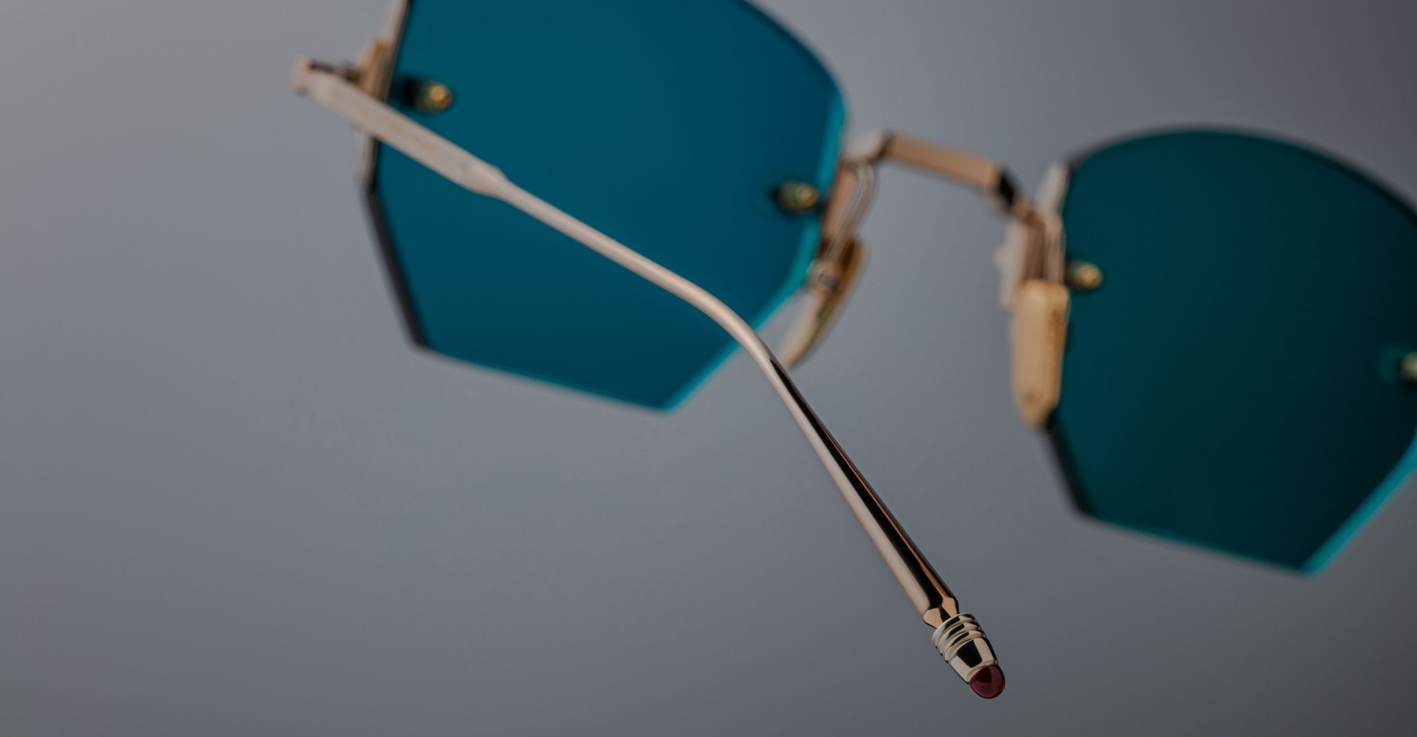 Close-up of Jacques Marie Mage Oatman gold-rimmed, blue-tinted hexagonal rimless sunglasses, highlighting the temple arm and handmade Japanese craftsmanship against a plain gray background.