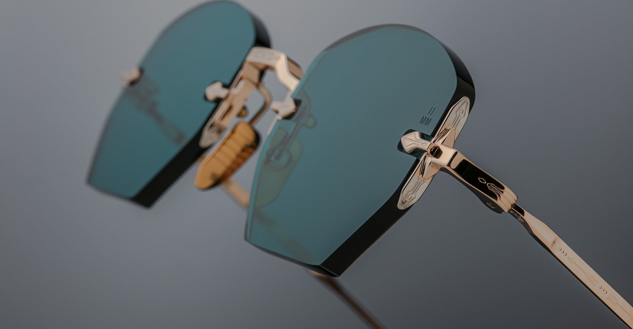Close-up of Jacques Marie Mage Oatman sunglasses by Jacques Marie Mage, featuring heart-shaped teal lenses and gold frames inspired by handmade Japanese eyewear artistry, set against a neutral background.