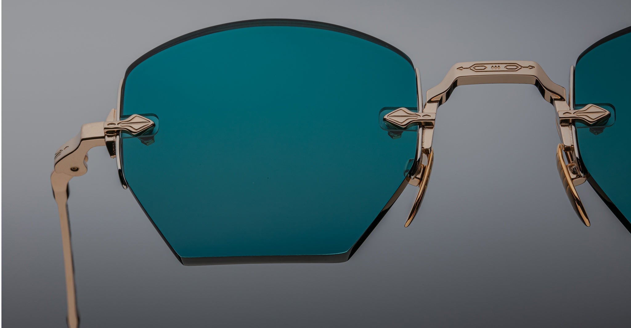 Close-up of Jacques Marie Mage Oatman hexagonal rimless sunglasses with a gold frame and large blue-green tinted lenses, highlighting the craftsmanship of handmade Japanese eyewear against a neutral background.