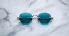 The Jacques Marie Mage Oatman sunglasses by Jacques Marie Mage feature round, blue-tinted lenses with silver frames, inspired by handmade Japanese eyewear, displayed on a white marble surface.