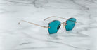 The Jacques Marie Mage Oatman, hexagonal rimless sunglasses with blue-tinted lenses and thin gold arms inspired by handmade Japanese eyewear, rests on a white marble surface.