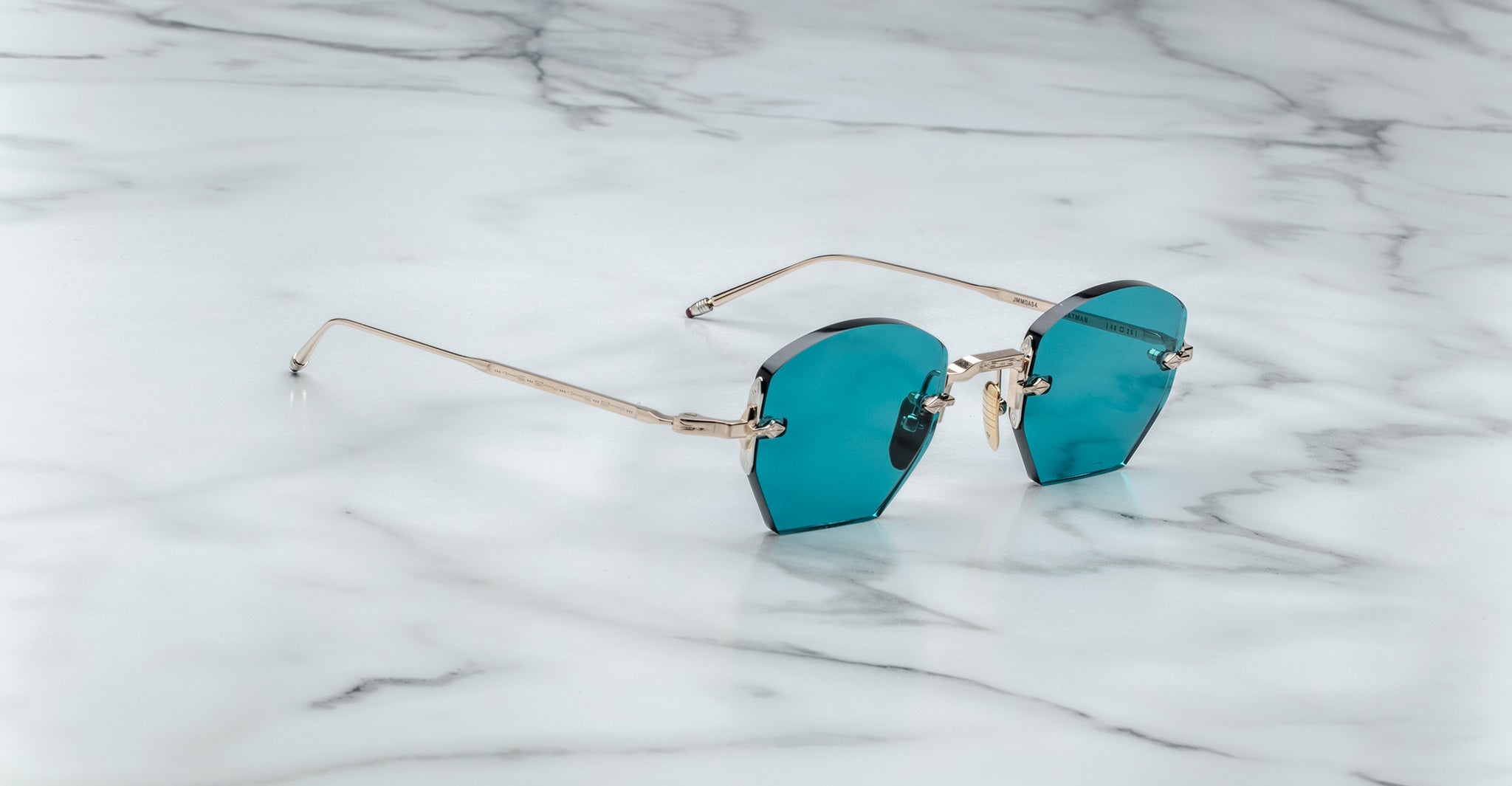 The Jacques Marie Mage Oatman, hexagonal rimless sunglasses with blue-tinted lenses and thin gold arms inspired by handmade Japanese eyewear, rests on a white marble surface.