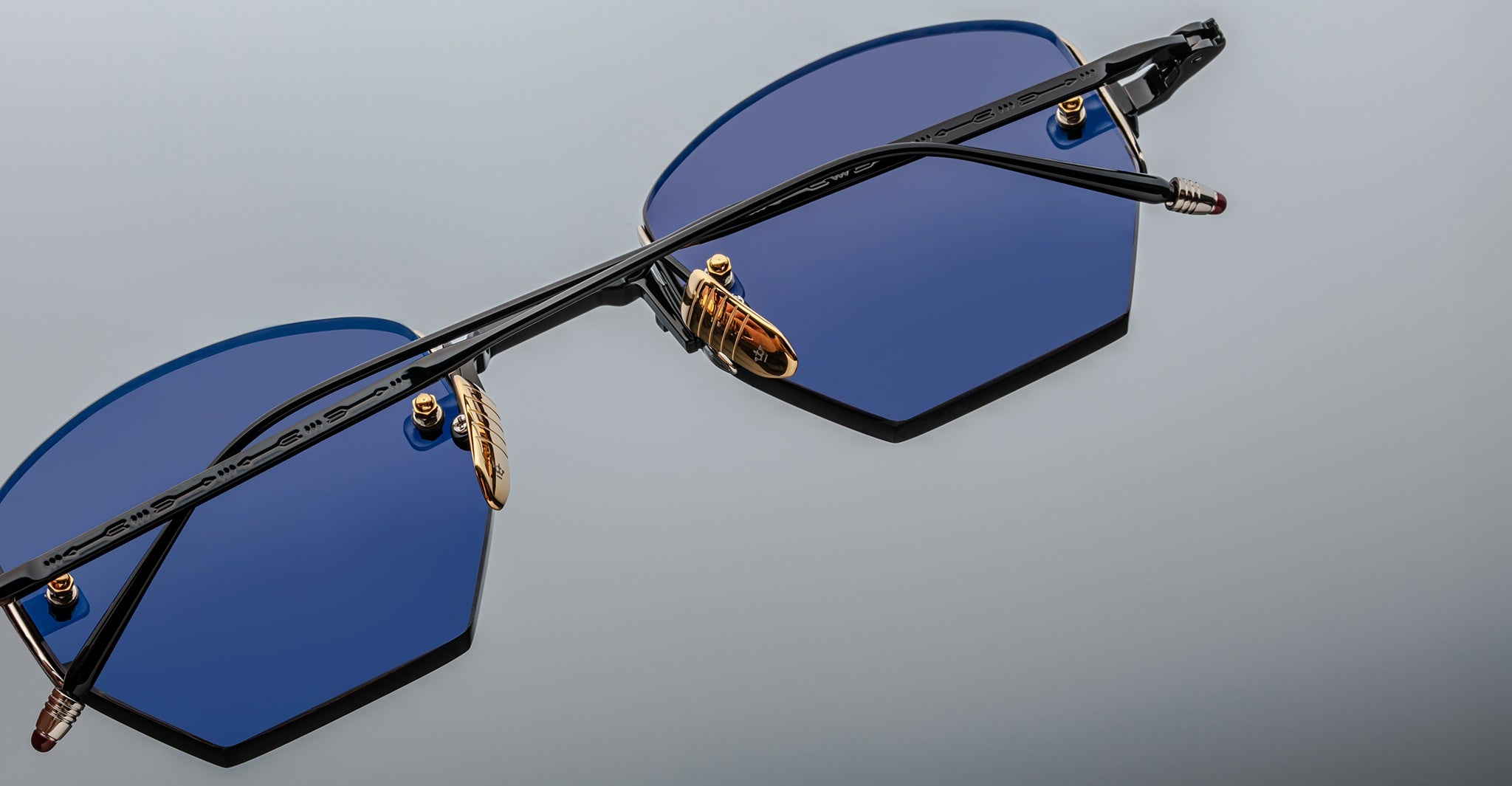 Close-up of Jacques Marie Mage Oatman hexagonal rimless sunglasses featuring geometric blue lenses, gold nose pads, and a black frame on a light gray background—showcasing Jacques Marie Mage’s exquisite handmade Japanese craftsmanship.