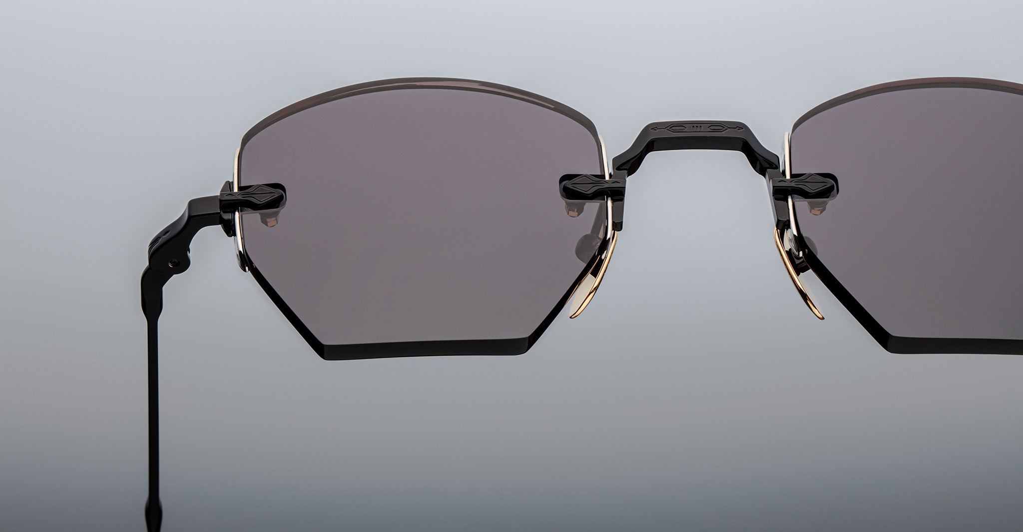 Close-up of Jacques Marie Mage Oatman sunglasses: hexagonal rimless frames with dark tinted lenses, black and gold metal hinges, and a minimalist design—showcasing handmade Japanese craftsmanship against a gradient background.