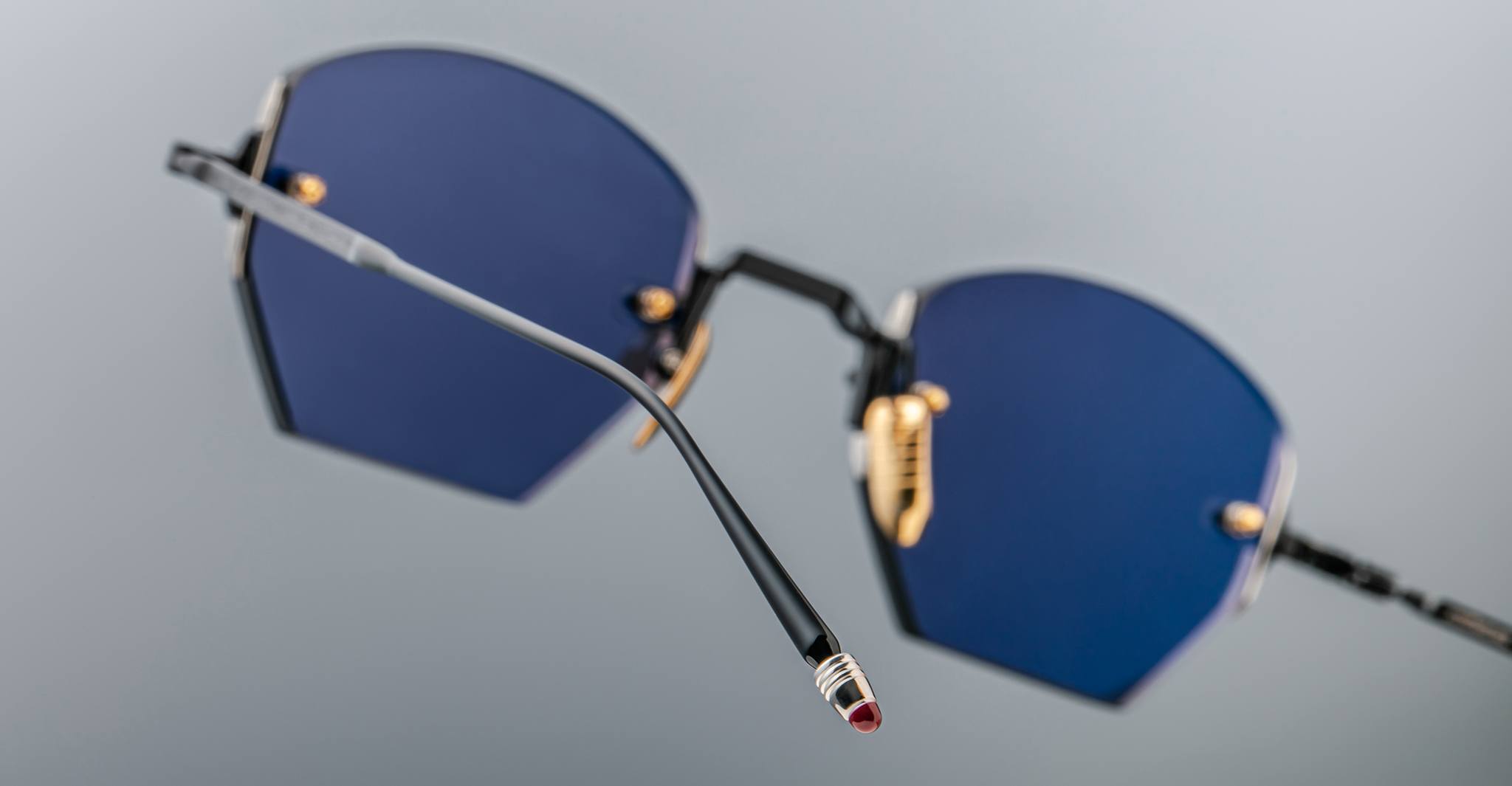 Close-up of Jacques Marie Mage Oatman sunglasses: hexagonal rimless design, blue-tinted lenses, gold accents, and black arms with metallic red tips—showcasing the exquisite handmade Japanese quality against a neutral background.