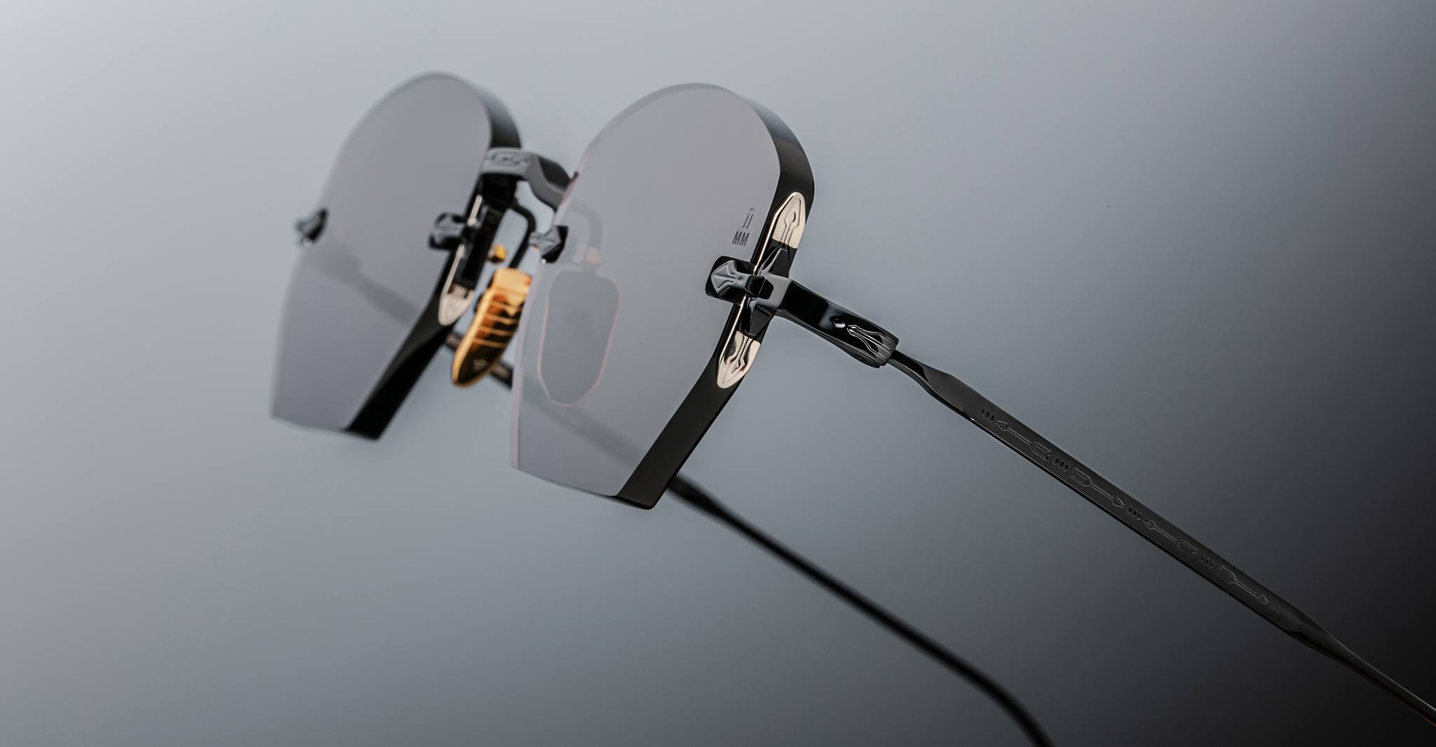 Close-up of Jacques Marie Mage Oatman: handmade Japanese hexagonal rimless sunglasses with dark polygonal lenses and thin black arms, shown against a grey background.