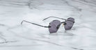 The Jacques Marie Mage Oatman, a pair of hexagonal rimless sunglasses with dark lenses and thin metal arms by Jacques Marie Mage, rests on a white marble surface.