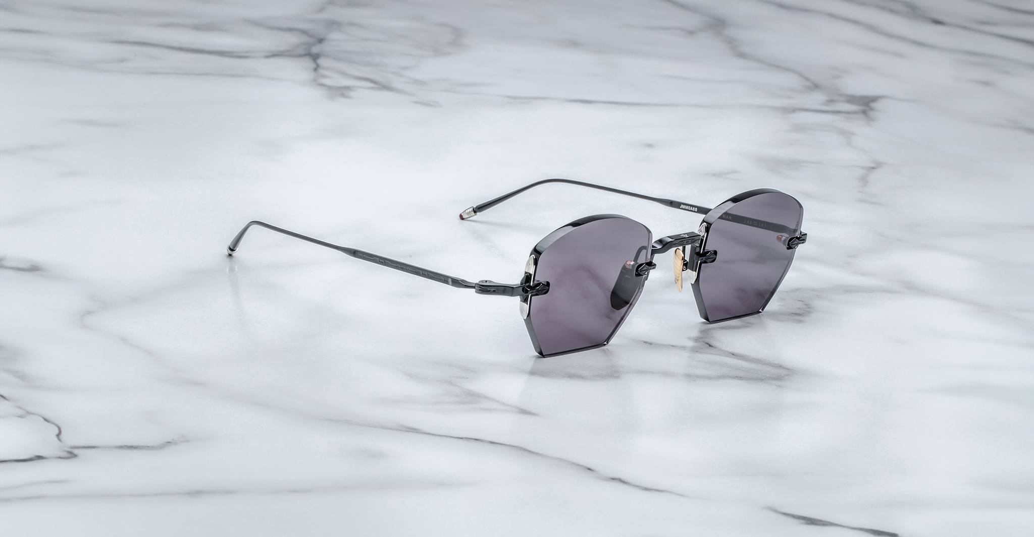 The Jacques Marie Mage Oatman, a pair of hexagonal rimless sunglasses with dark lenses and thin metal arms by Jacques Marie Mage, rests on a white marble surface.