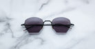 A pair of Jacques Marie Mage Oatman hexagonal rimless sunglasses with dark lenses rests on a white marble surface.