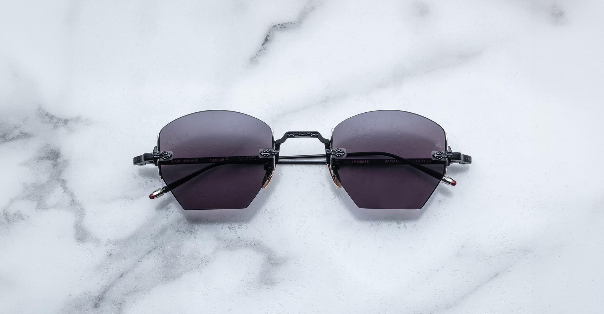 A pair of Jacques Marie Mage Oatman hexagonal rimless sunglasses with dark lenses rests on a white marble surface.