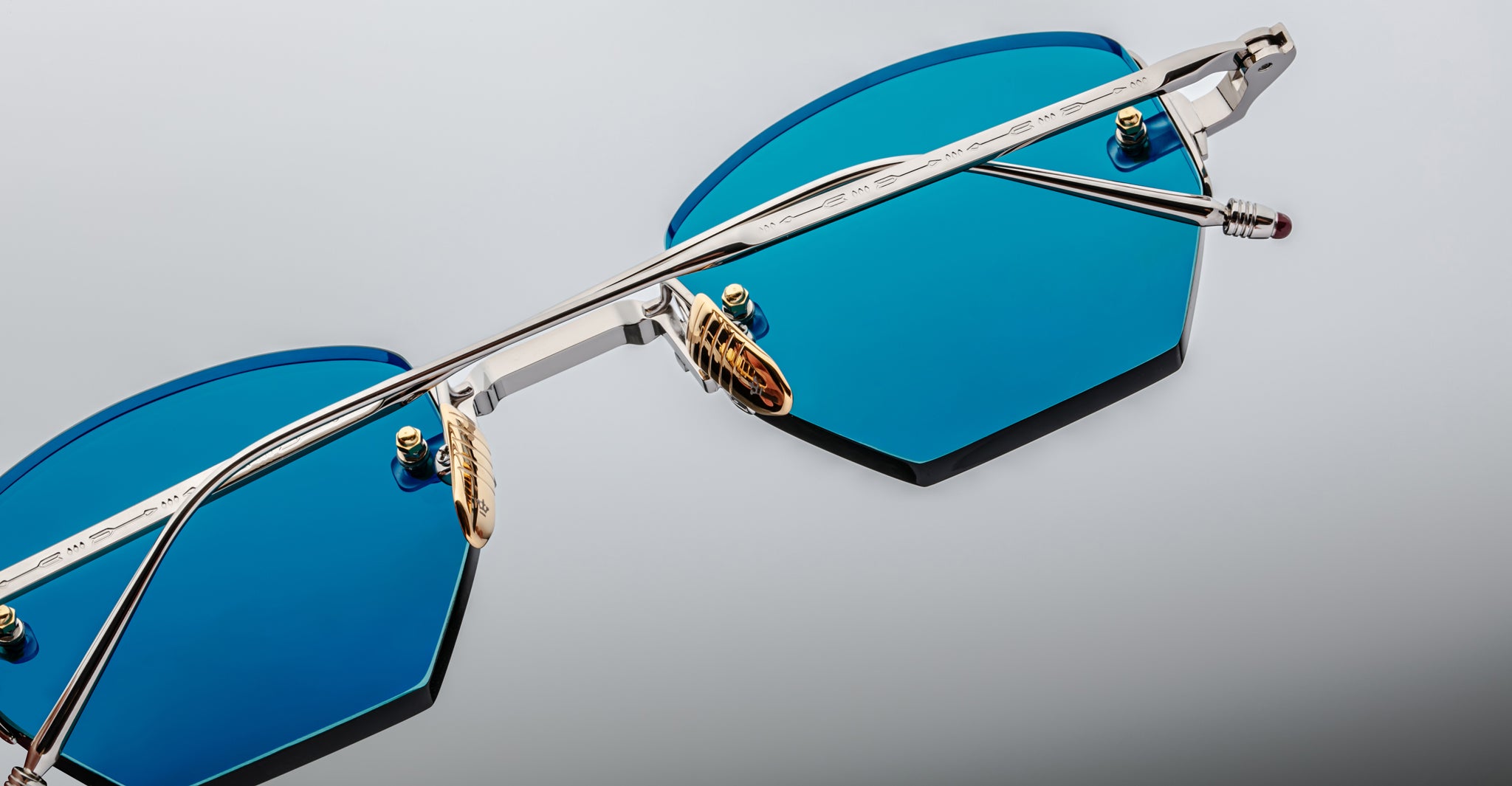 Close-up of Jacques Marie Mage Oatman hexagonal rimless sunglasses with blue lenses, silver metal frame, and gold nose pads—handmade Japanese eyewear by Jacques Marie Mage—set against a light background.