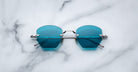 A pair of Jacques Marie Mage Oatman hexagonal rimless sunglasses with blue-tinted geometric lenses and metal frames rests on a white marble surface.