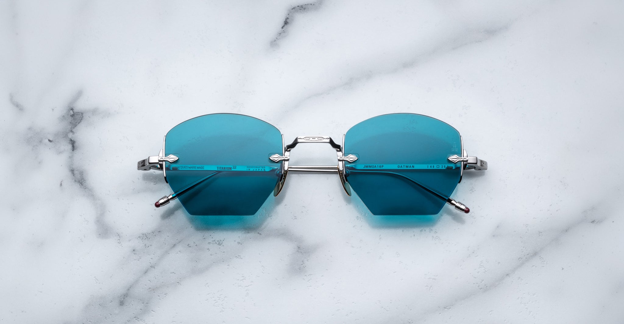 A pair of Jacques Marie Mage Oatman hexagonal rimless sunglasses with blue-tinted geometric lenses and metal frames rests on a white marble surface.