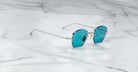 The Jacques Marie Mage Oatman, a pair of hexagonal rimless sunglasses with blue-tinted lenses and thin silver arms, is displayed on a white marble surface, highlighting the elegance of handmade Japanese eyewear.