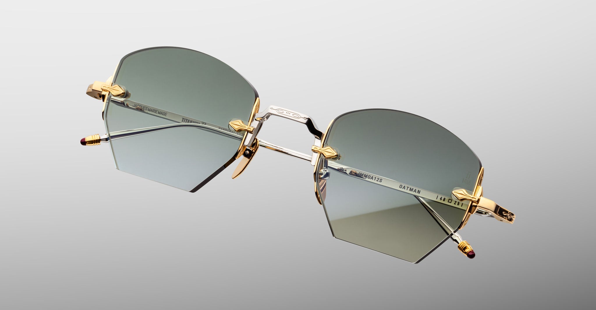 The Jacques Marie Mage Oatman sunglasses feature a geometric hexagonal rimless design with gold accents, gray-green lenses, and slim metal arms. Inspired by handmade Japanese craftsmanship, shown on a light gray background.