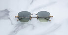 A pair of Jacques Marie Mage Oatman hexagonal rimless sunglasses with geometric gray lenses and gold metal temples rests on a white marble surface.
