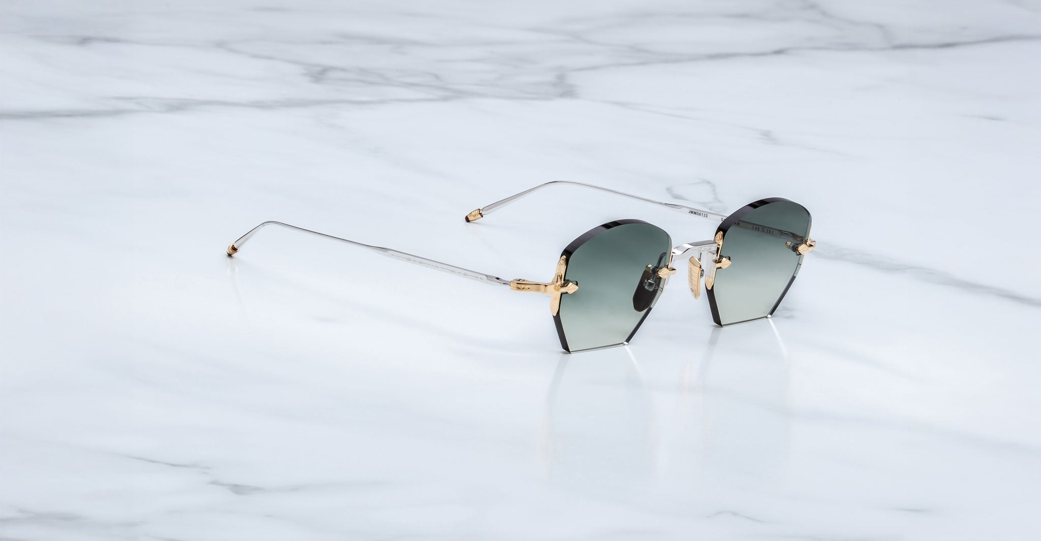 A pair of Jacques Marie Mage Oatman hexagonal rimless sunglasses with gold temples and dark lenses sits on a white marble surface.