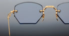 Close-up of Jacques Marie Mage Oatman eyeglasses by Jacques Marie Mage, featuring gold rims and blue-tinted geometric lenses, inspired by handmade Japanese eyewear and set against a neutral gradient background.