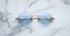 The Jacques Marie Mage Oatman sunglasses, featuring gold hexagonal frames and light blue lenses, are displayed on a white marble surface.