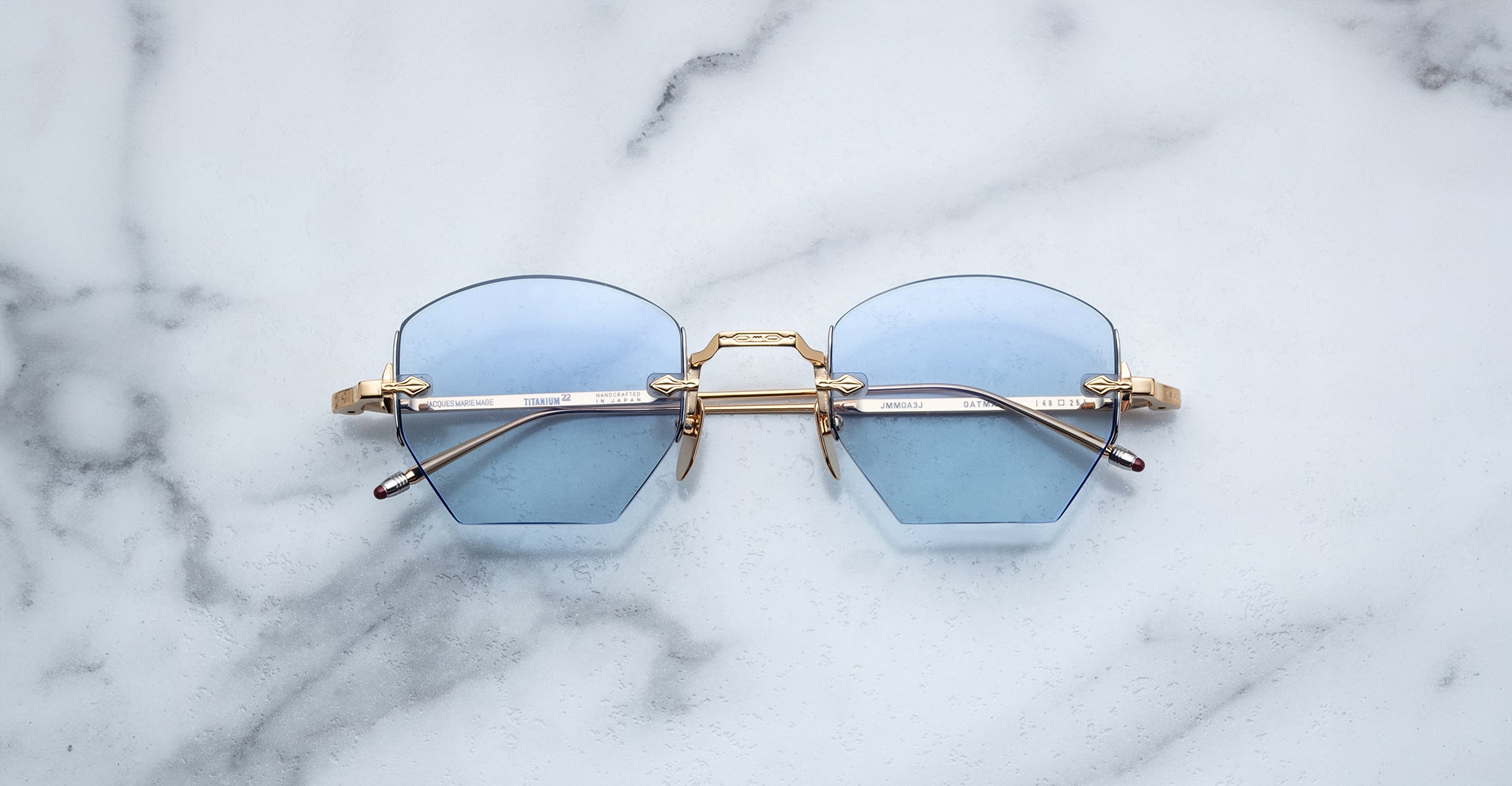 The Jacques Marie Mage Oatman sunglasses, featuring gold hexagonal frames and light blue lenses, are displayed on a white marble surface.