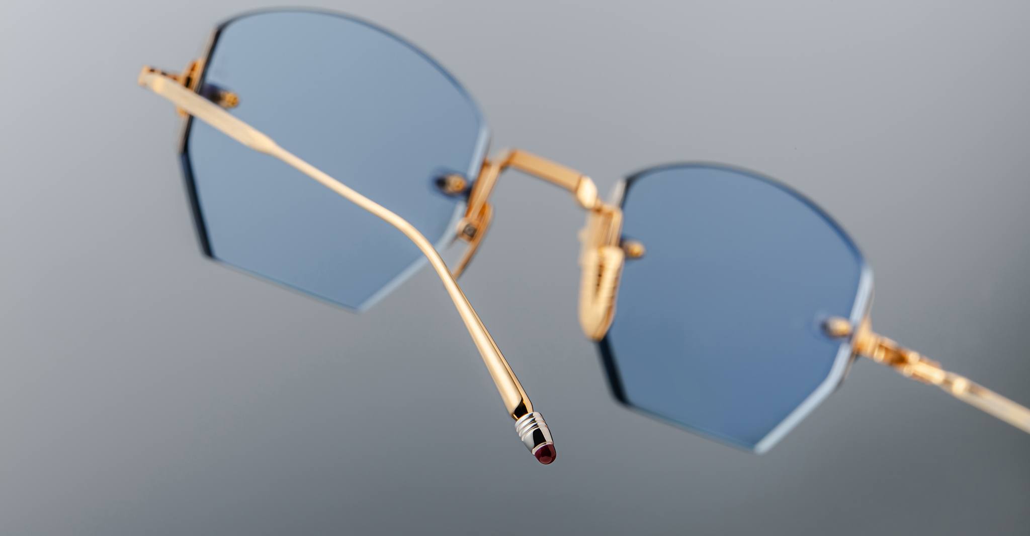 Close-up of Jacques Marie Mage Oatman gold-framed handmade Japanese eyewear featuring blue hexagonal lenses, with a focus on the temple arm against a plain gray background.