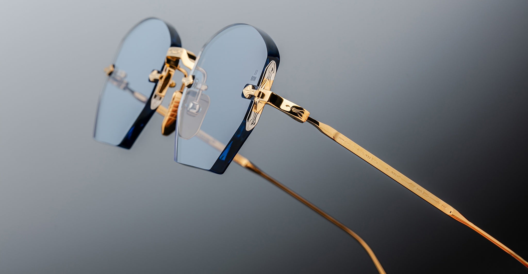 A close-up of Jacques Marie Mage Oatman hexagonal rimless sunglasses, featuring blue-tinted lenses and gold metal temples by Jacques Marie Mage, set against a gradient gray background.