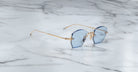 Jacques Marie Mage Oatman sunglasses feature a hexagonal rimless design with blue-tinted lenses and gold metal arms, inspired by handmade Japanese eyewear, displayed on a white marble surface.