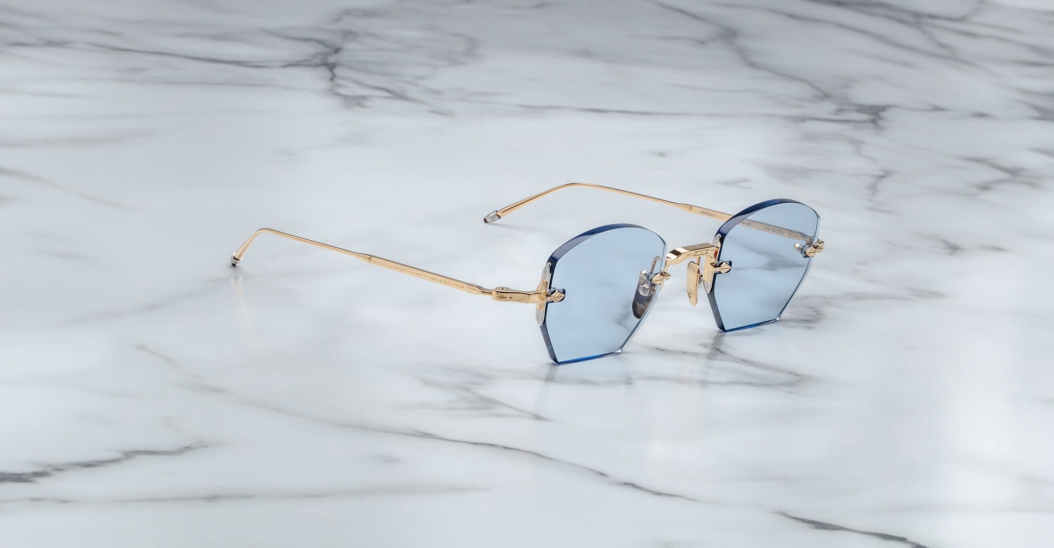 Jacques Marie Mage Oatman sunglasses feature a hexagonal rimless design with blue-tinted lenses and gold metal arms, inspired by handmade Japanese eyewear, displayed on a white marble surface.