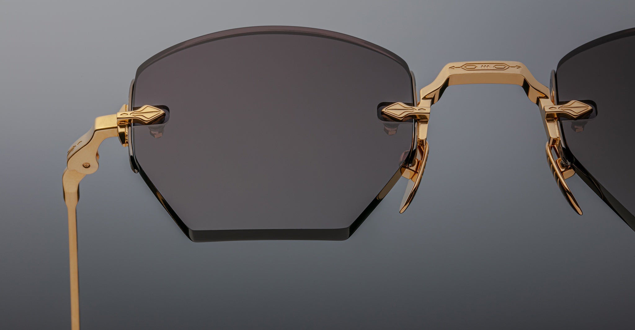 Close-up of Jacques Marie Mage Oatman sunglasses: luxury hexagonal rimless dark lenses with a gold metal frame, detailed hinge, and geometric design. Exquisite handmade Japanese eyewear by Jacques Marie Mage against a plain background.