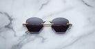 The Jacques Marie Mage Oatman hexagonal rimless sunglasses, featuring gold temples and dark lenses, rest on a white marble surface—highlighting the brand's exquisite handmade Japanese eyewear craftsmanship.