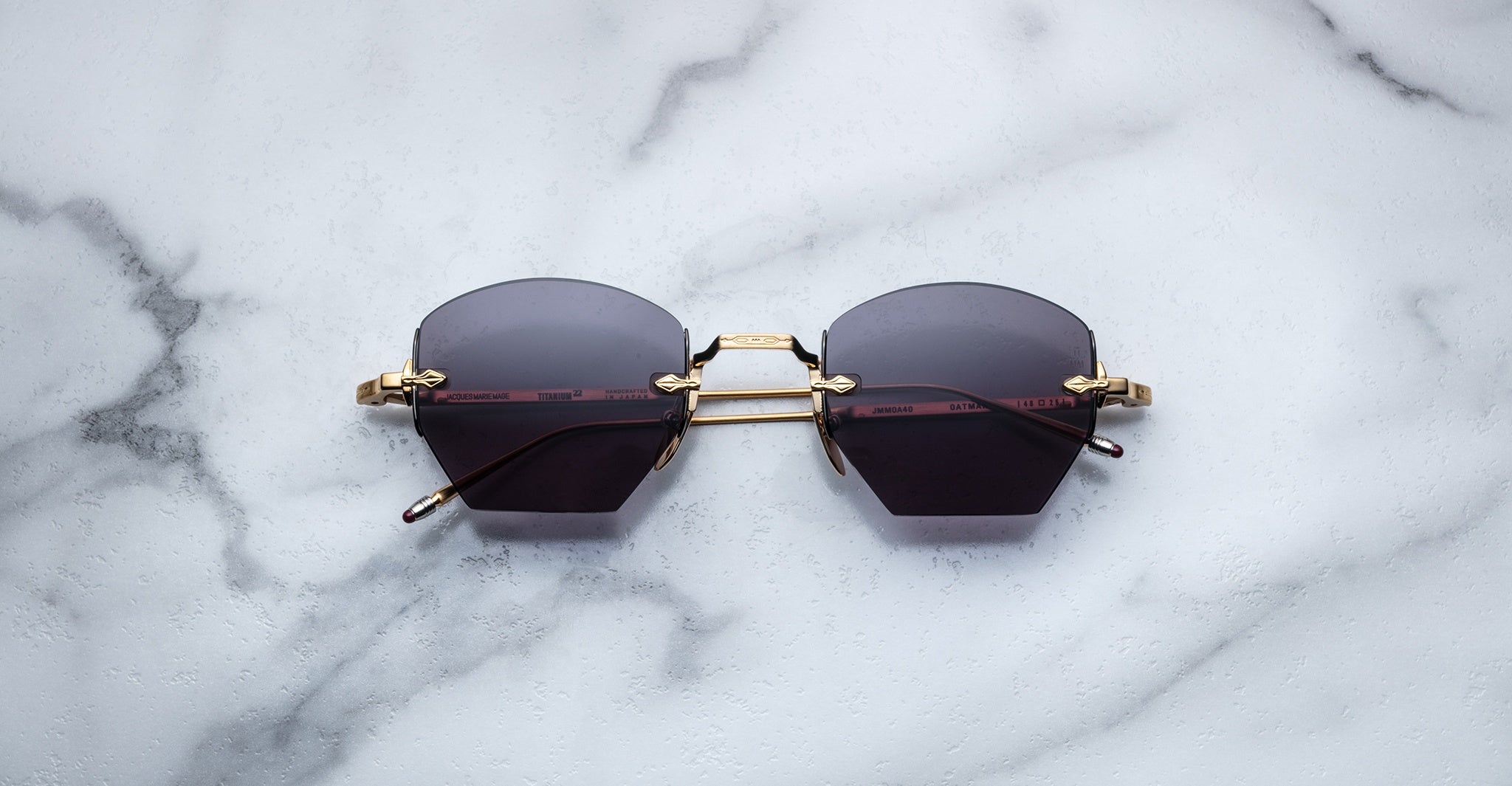 The Jacques Marie Mage Oatman hexagonal rimless sunglasses, featuring gold temples and dark lenses, rest on a white marble surface—highlighting the brand's exquisite handmade Japanese eyewear craftsmanship.