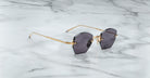 The Jacques Marie Mage Oatman sunglasses, featuring hexagonal rimless dark lenses and gold metal arms, rest elegantly on a white marble surface.