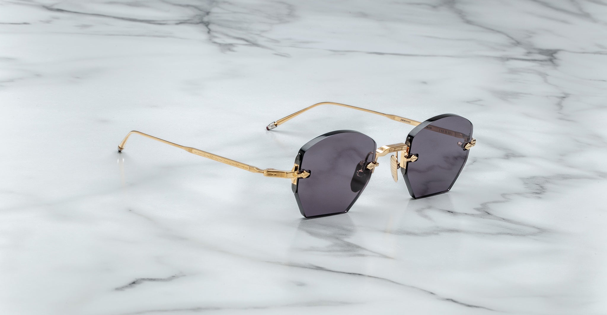 The Jacques Marie Mage Oatman sunglasses, featuring hexagonal rimless dark lenses and gold metal arms, rest elegantly on a white marble surface.