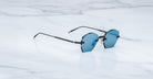 The Jacques Marie Mage Oatman, handmade Japanese hexagonal rimless sunglasses with blue-tinted lenses and thin black arms, is displayed on a white marble surface.