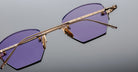 Close-up of Jacques Marie Mage Oatman eyewear: gold-framed, hexagonal rimless sunglasses with striking purple lenses by Jacques Marie Mage, set against a grey background.