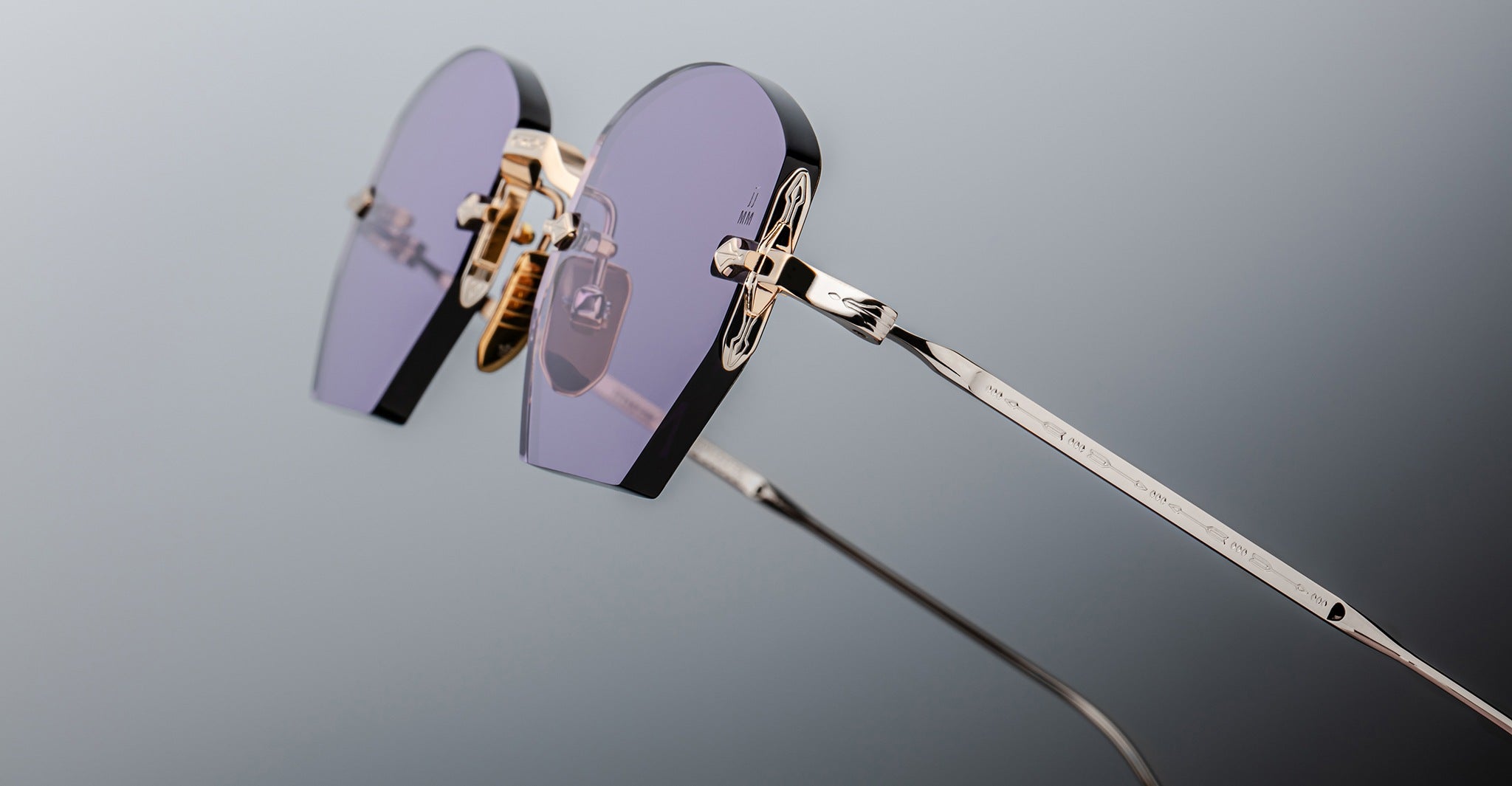 Close-up of Jacques Marie Mage Oatman sunglasses: stylish hexagonal rimless frames with heart-shaped purple lenses, gold nose pads, and thin metal arms, evoking handmade Japanese eyewear, set against a neutral gray background.