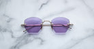 Jacques Marie Mage Oatman sunglasses by Jacques Marie Mage feature hexagonal rimless frames, purple lenses, and silver arms inspired by Japanese craftsmanship, shown elegantly on a white marble surface.