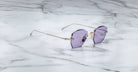 A pair of Jacques Marie Mage Oatman sunglasses, featuring hexagonal rimless frames, purple-tinted lenses, and thin gold temples, rests on a white marble surface.