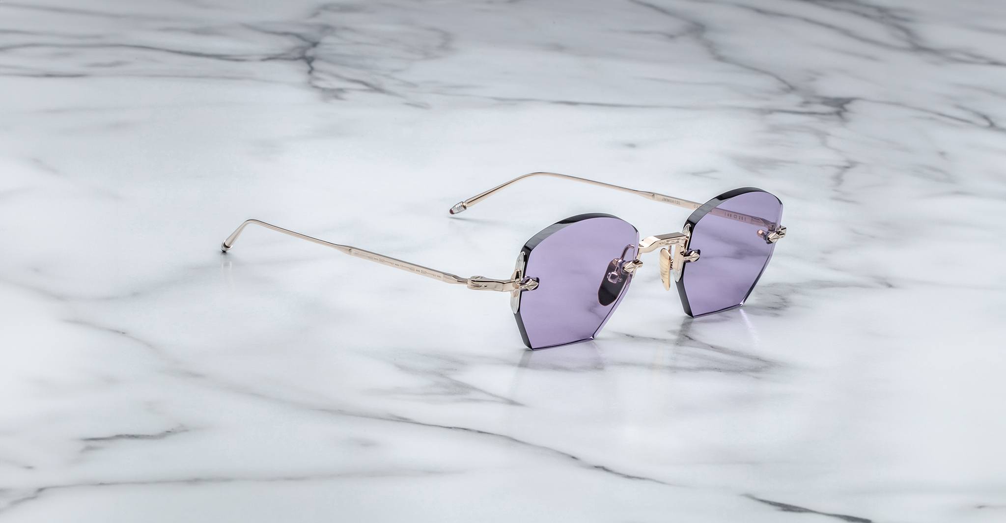 A pair of Jacques Marie Mage Oatman sunglasses, featuring hexagonal rimless frames, purple-tinted lenses, and thin gold temples, rests on a white marble surface.