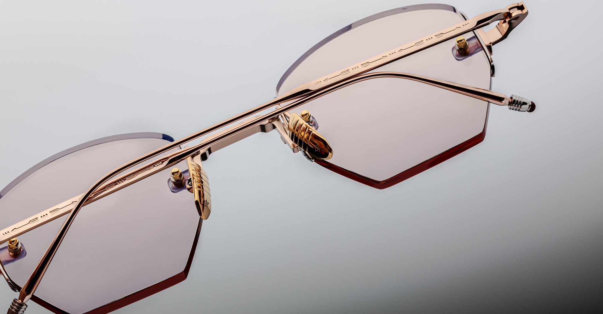 Close-up of Jacques Marie Mage Oatman: handmade Japanese hexagonal rimless sunglasses with gold-colored frames and light-tinted lenses, shown against a neutral background.