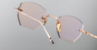 A close-up of Jacques Marie Mage Oatman gold-framed, hexagonal rimless sunglasses with pink-tinted lenses highlights the refined artistry of handmade Japanese eyewear by Jacques Marie Mage against a light gray background.