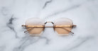 The Jacques Marie Mage Oatman sunglasses feature hexagonal rimless gold frames and light brown gradient lenses, inspired by Jacques Marie Mage's handmade Japanese eyewear, shown on a white marble surface.