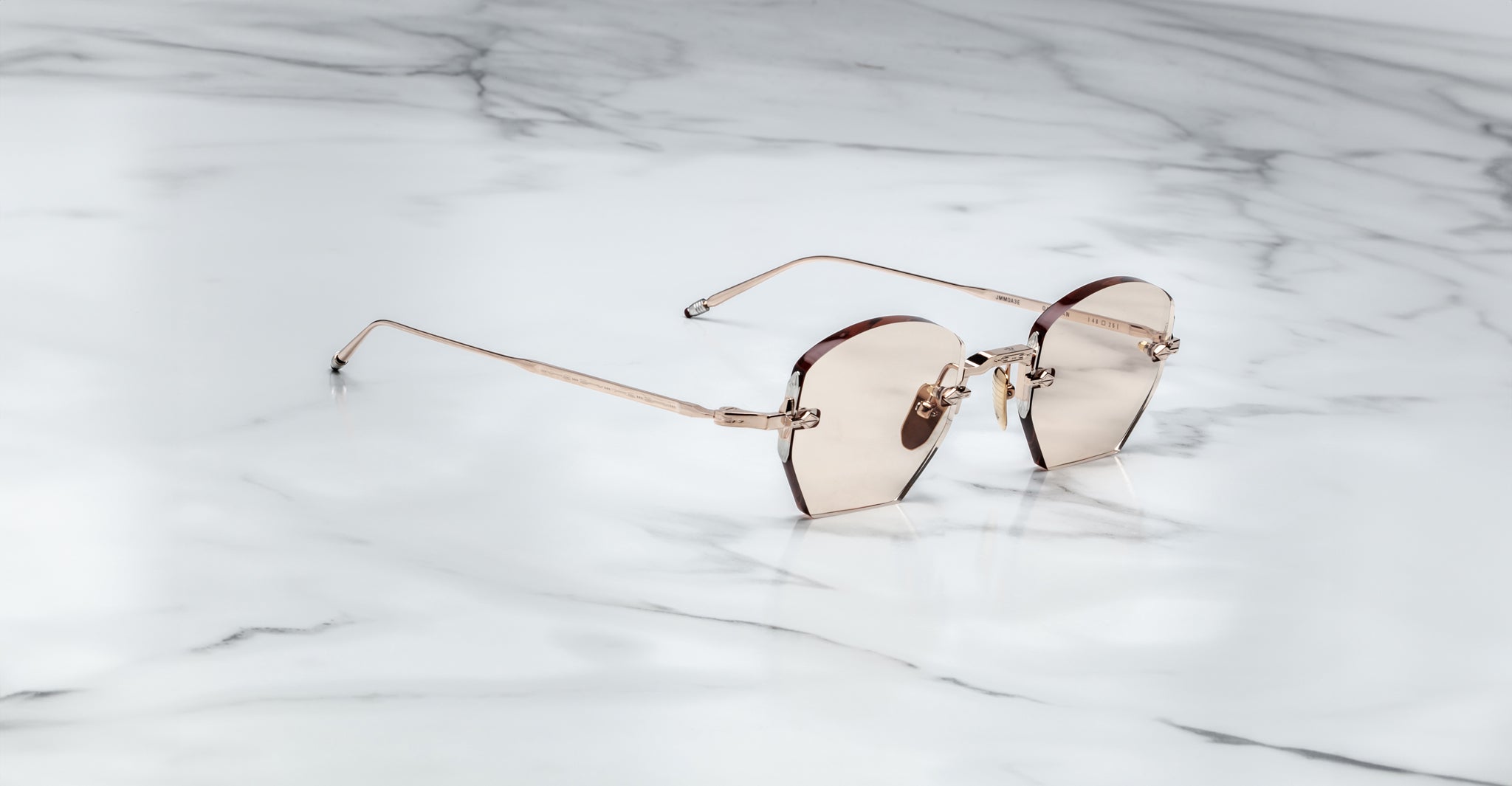 The Jacques Marie Mage Oatman, hexagonal rimless sunglasses with rose-tinted lenses and thin metal arms inspired by Japanese craftsmanship, rest elegantly on a white marble surface.